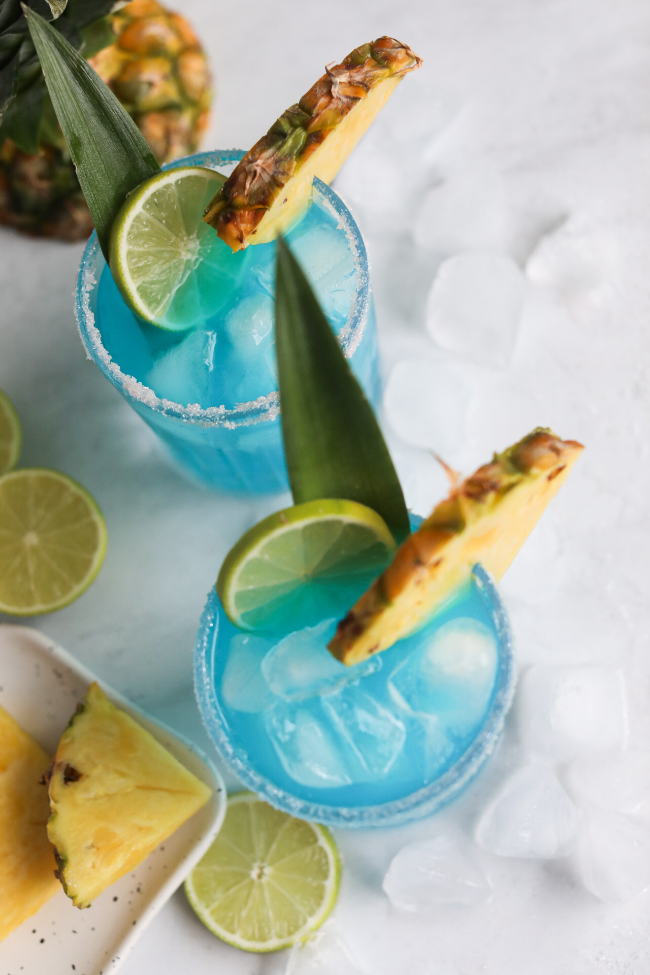 Top down view of blue margarita showing the top of two glasses filled with margarita cocktail. Sliced pineapple, lime and ice cubes added for styling purposes. 