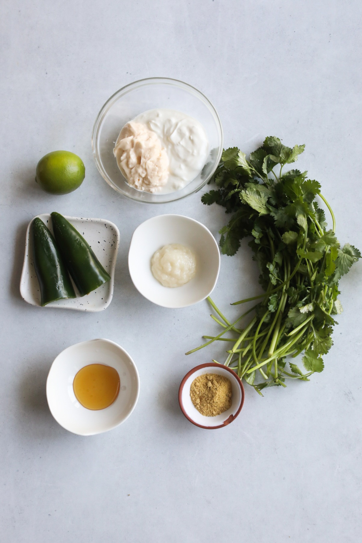 Cilantro lime sauce ingredients in a flat lay. Small white bowls, clear bowl and small white plate with mayo, greek yogurt, jalapeño, garlic paste, honey, chicken bouillon, cilantro and lime.