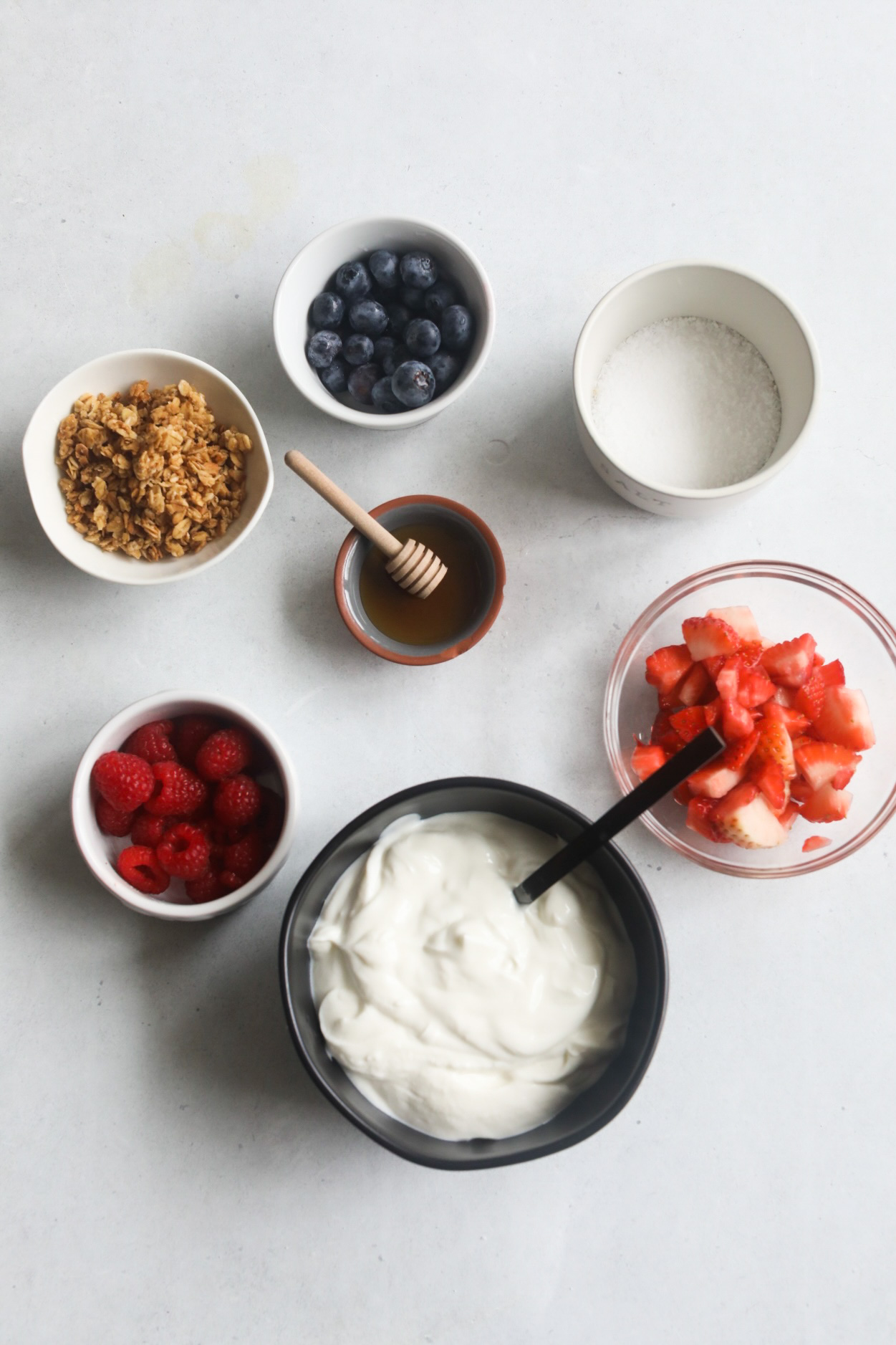 Greek Yogurt Parfait ingredient image with flat of small white, clear and black bowls. Greek yogurt, raspberries, strawberries, granola, blueberries, salt and honey with honey stick.