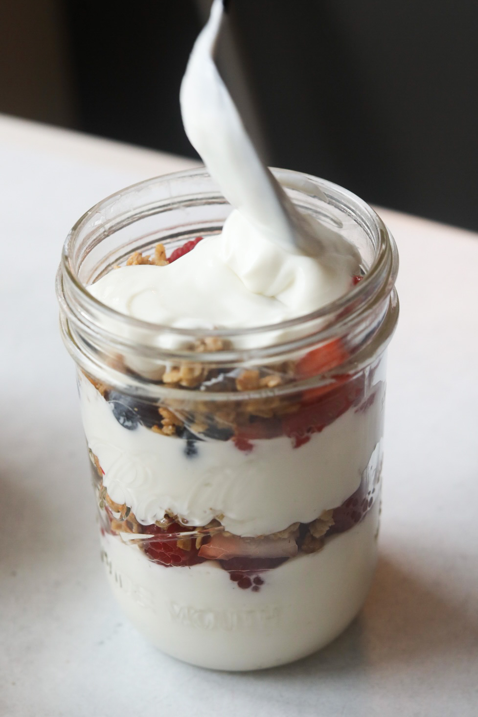 Greek yogurt being spooned into a short glass mason jar.