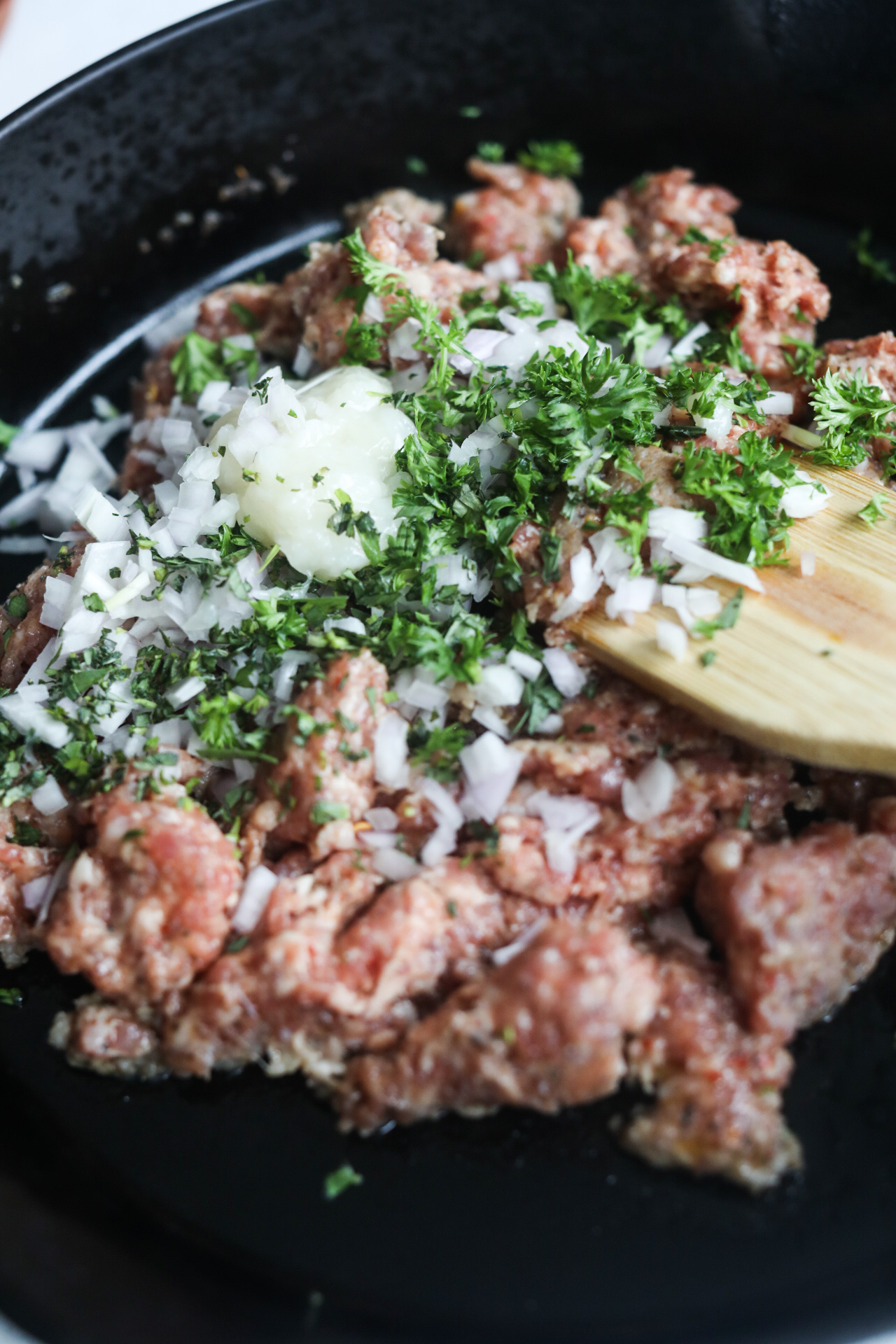 how to make one pot lasagna Par cooked ground beef and pork mixture in a black cast iron skillet topped with garlic paste, onion and fresh parsley. Wooden spoon is mixing ingredients.