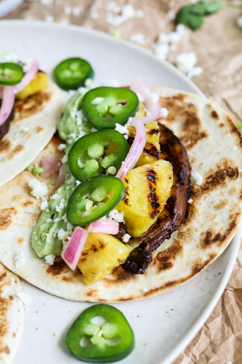 Portobello, mushroom tacos plated with grilled pineapple, jalapeño, pickled onion and avocado cream on a white plate.