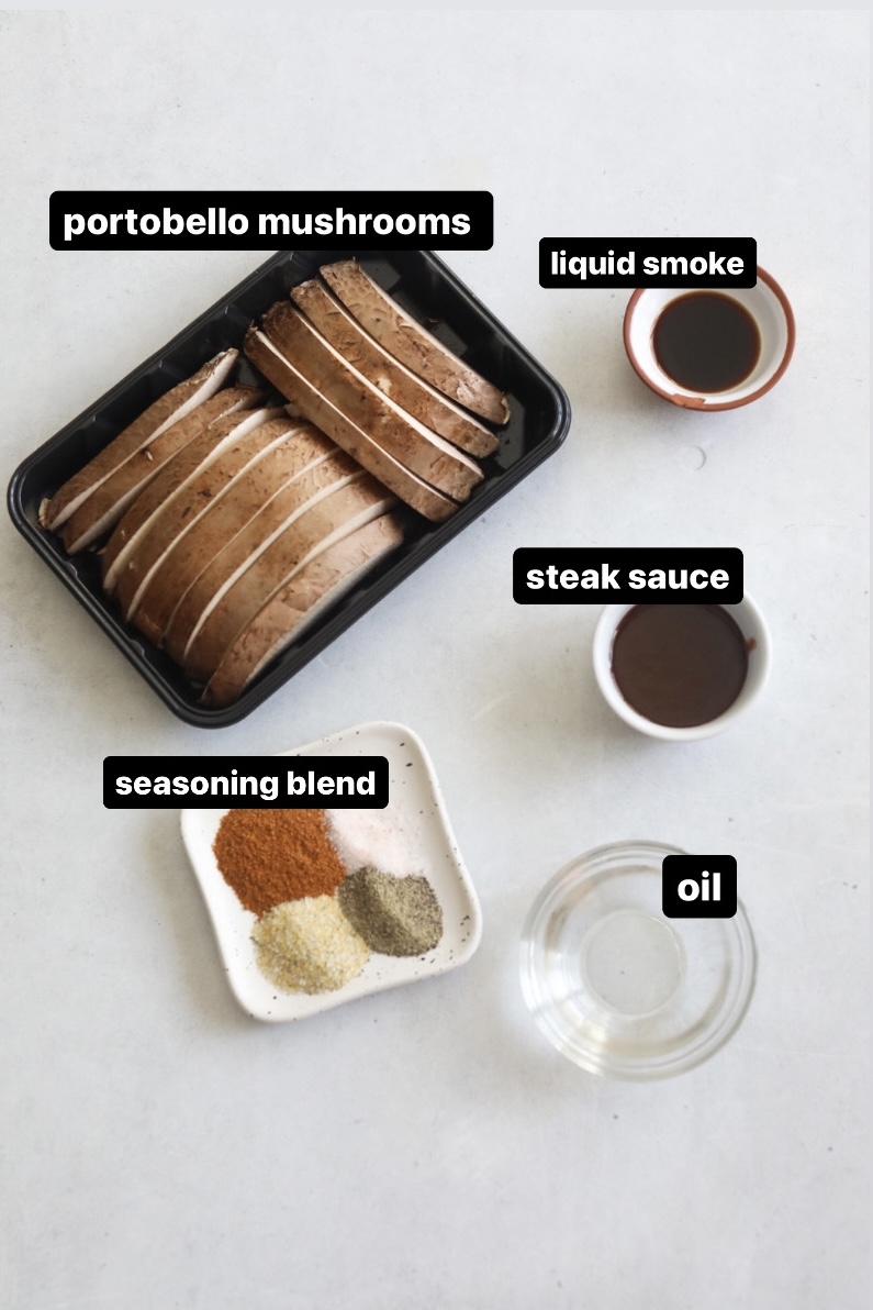 Mushroom tacos ingredients in a flat lay. Portobello mushrooms on a black tray, small white plate with seasoning blend, small bowls of liquid smoke, water and steak sauce.