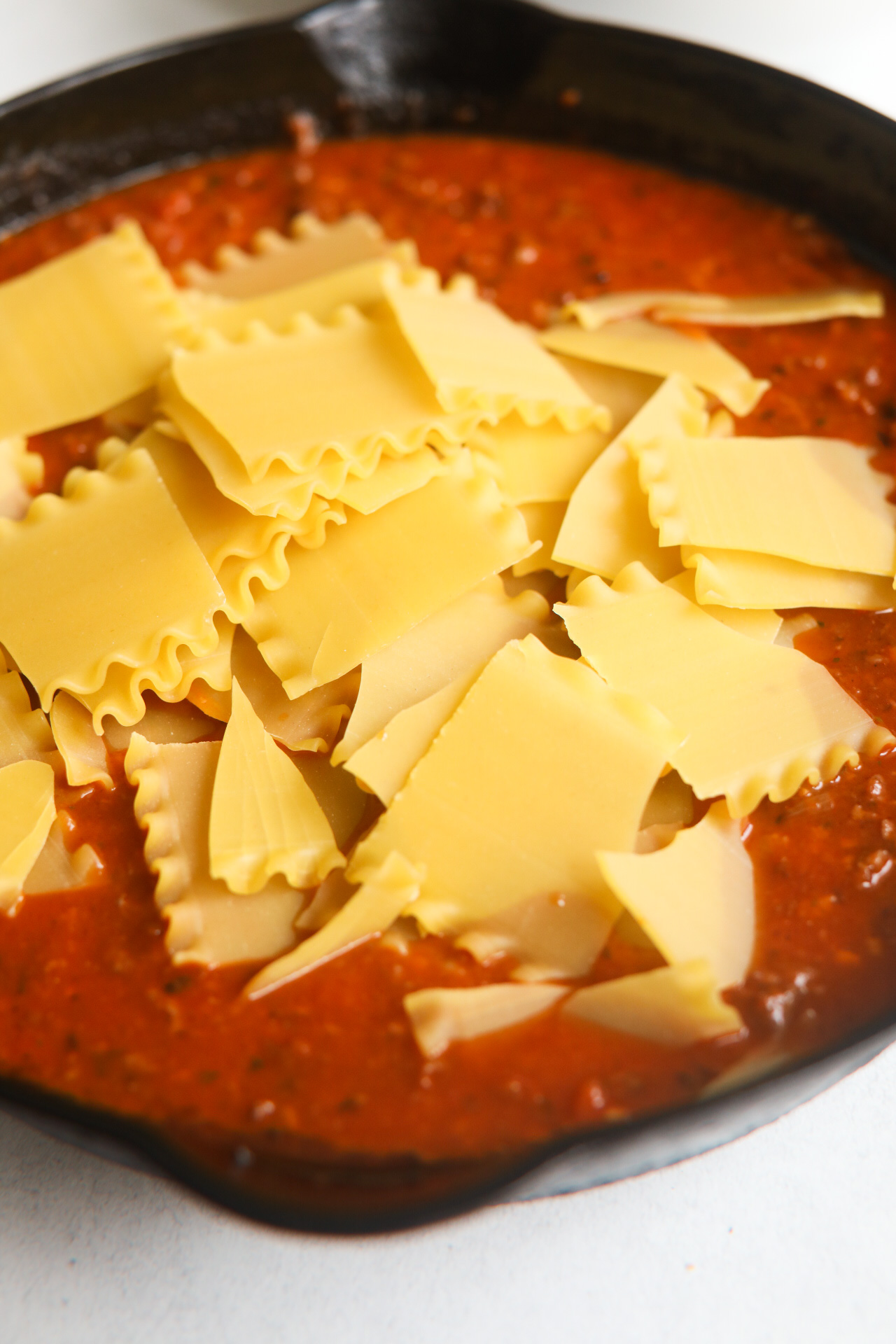 Process shot with black cast iron skillet filled with cooked ground beef and pork mixture topped with beef broth, marinara sauce and broken uncooked lasagna noodles.