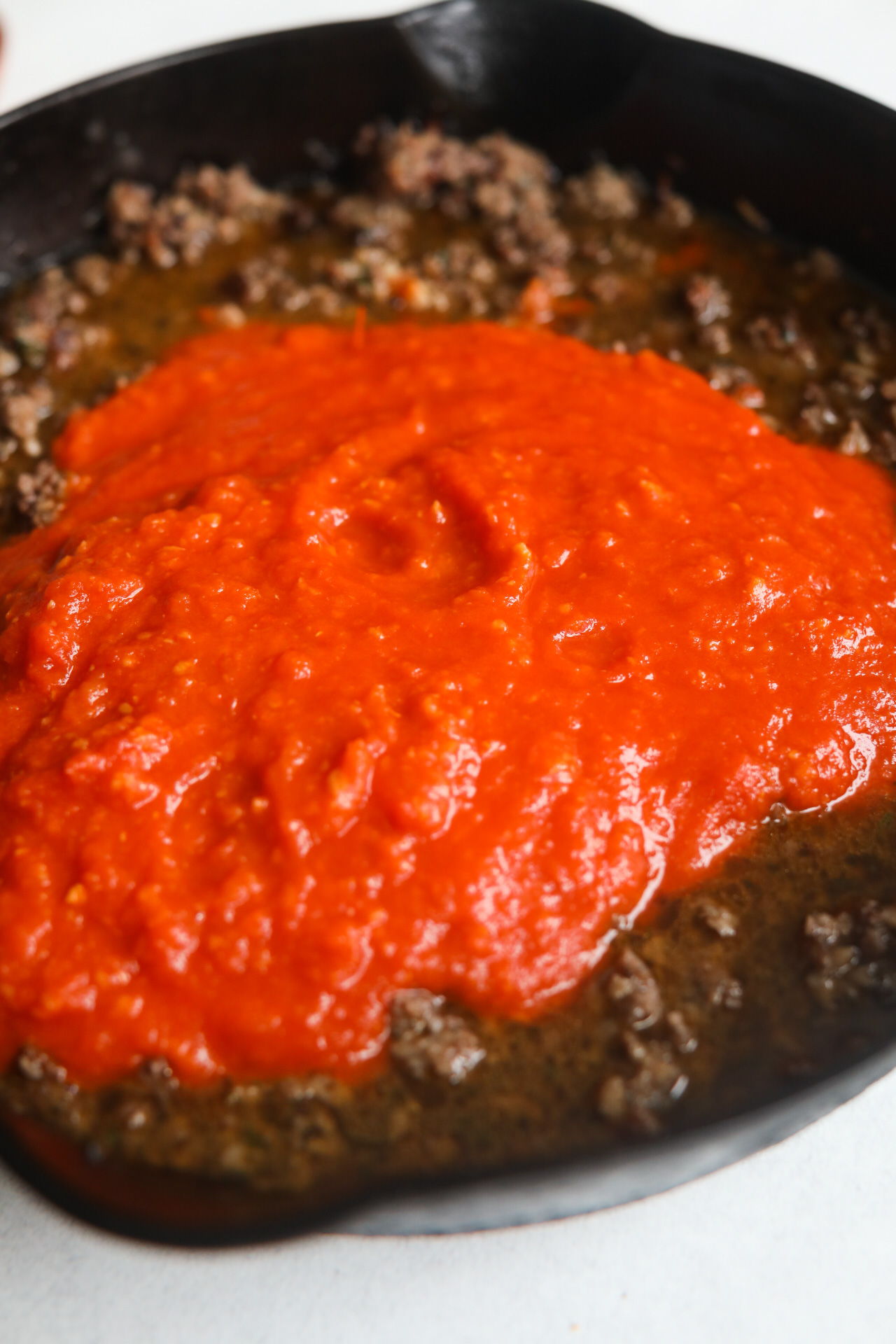 Process shot with black cast iron skillet filled with cooked ground beef and pork mixture topped with beef broth and marinara sauce.