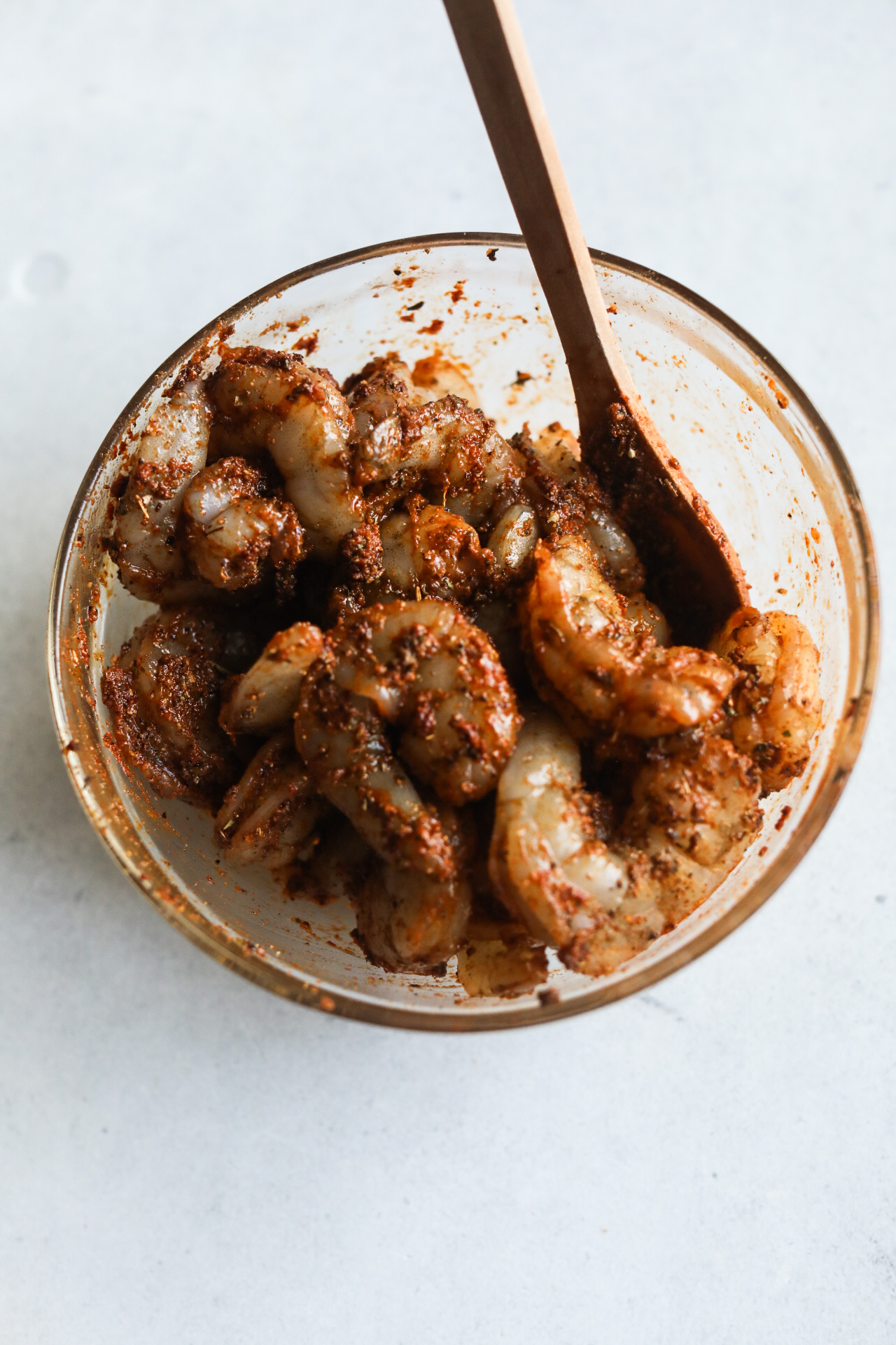 Shrimp marinated with blackening seasoning in a small clear bowl with wooden spoon.