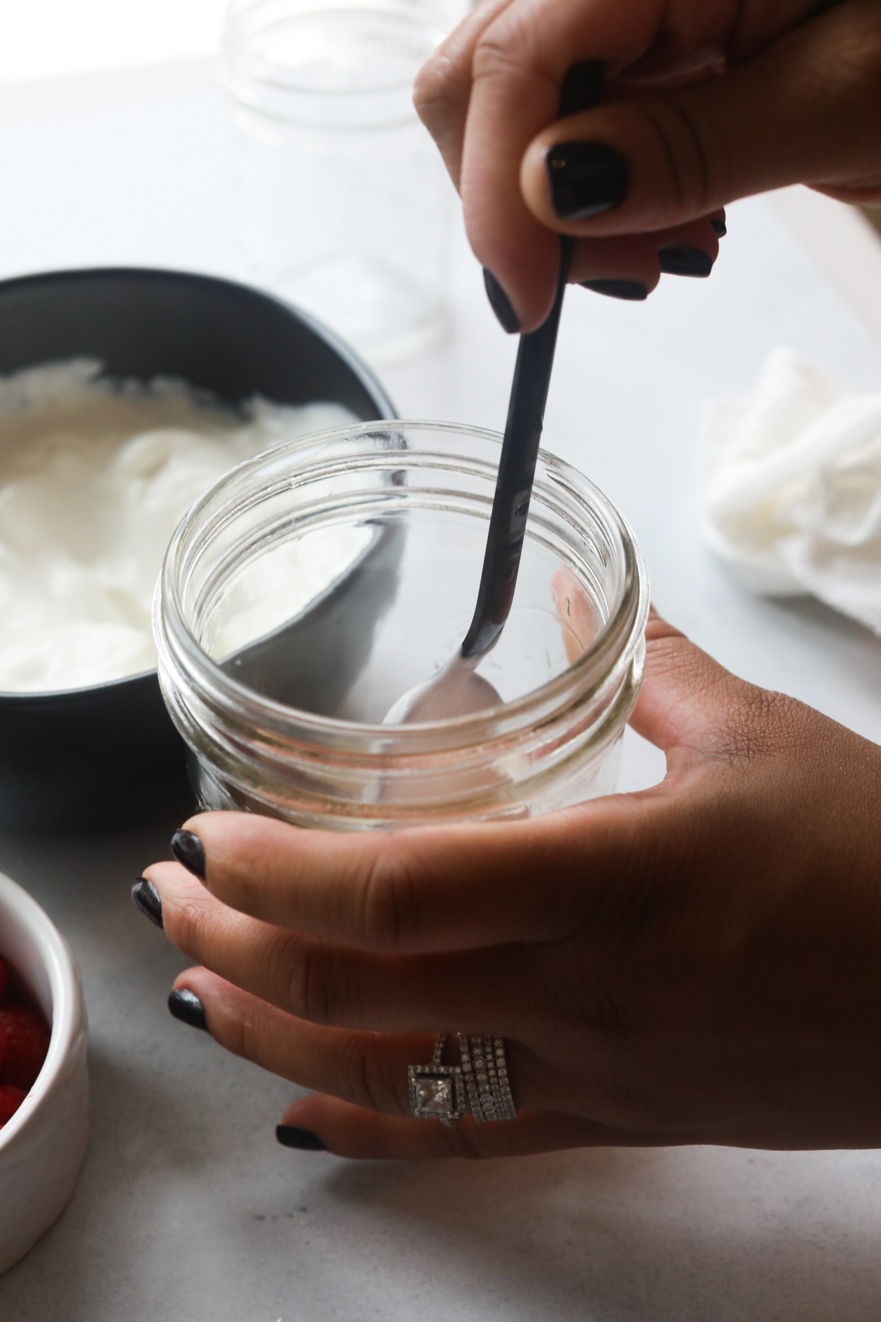 Preparing Greek Yogurt Parfait into a short glass mason jar with hands adding yogurt with a black spoon.
