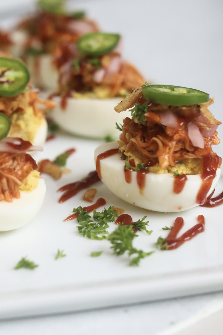Deviled eggs with BBQ chicken, topped with pickled red onion and sliced jalapeño. Deviled eggs are garnished with fresh parsley, fried onions and bbq sauce drizzle. 