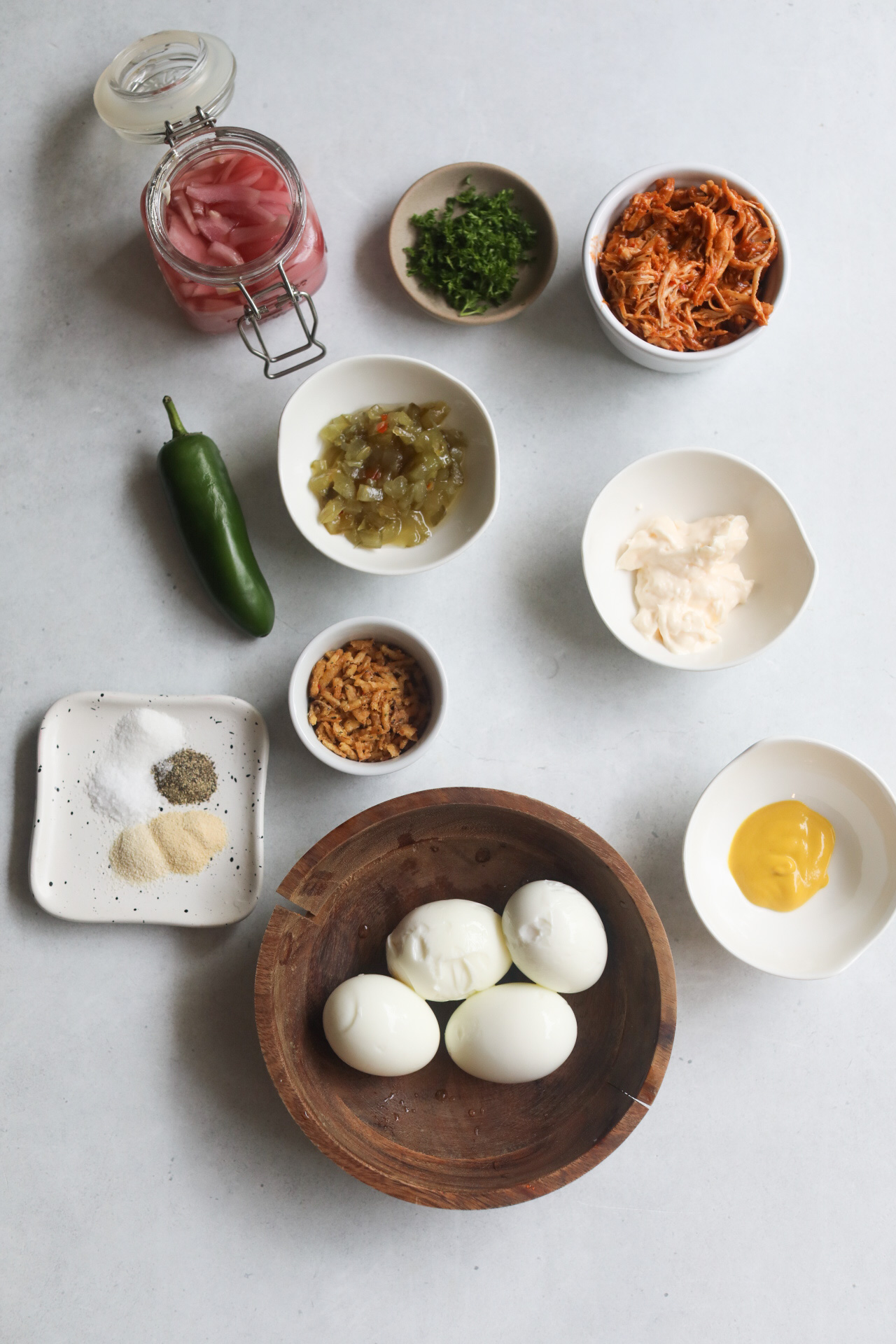 Loaded deviled eggs ingredients in a flat lay. In small bowls, shredded BBQ chicken, pickled red onion, mayo, mustard, relish and seasonings.