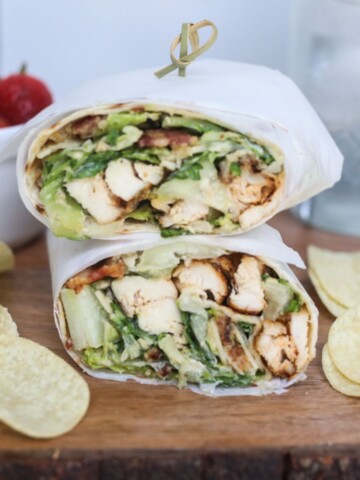 Featured image of Grilled Chicken Caesar Wrap. Wrap is served on wooden board with chips and cup of strawberries out of focus for styling purposes.