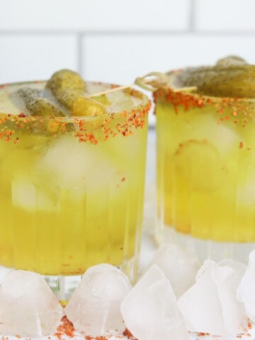 Featured Image for pickle margarita. Two short glasses filled with green margarita mix, topped with gherkin style pickles.