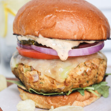 Tuna burger plated and stacked with brioche buns, slathered with remoulade sauce, tuna patty with havarti cheese, sliced tomato, red onion, arugula and top bun. Chips are added for styling purposes.