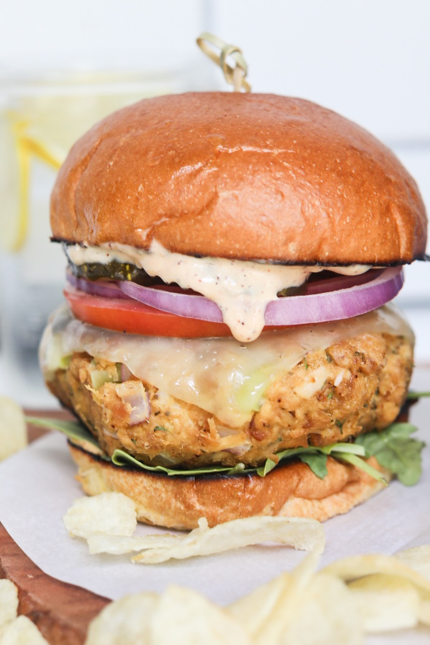 Tuna burger plated and stacked with brioche buns, slathered with remoulade sauce, tuna patty with havarti cheese, sliced tomato, red onion, arugula and top bun. Chips are added for styling purposes. 