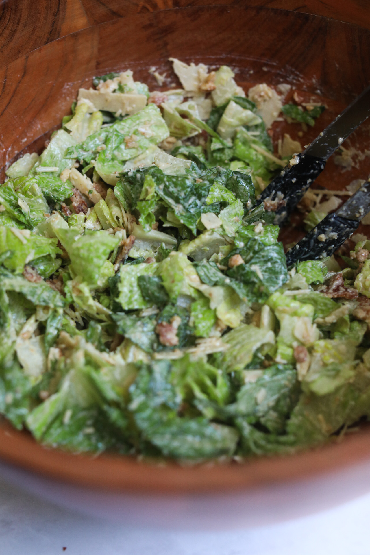 Process shot of caesar salad with romaine lettuce, parmesan cheese and bacon in a large brown wooden bowl, mixed with caesar dressing.