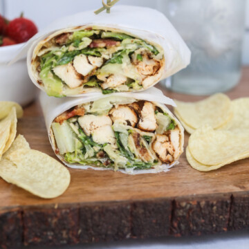 Grilled Chicken Caesar Wrap final image showing completed recipe. Caesar wrap is wrapped in wax paper, stacked and chips and a cup of strawberries are added for styling purposes.