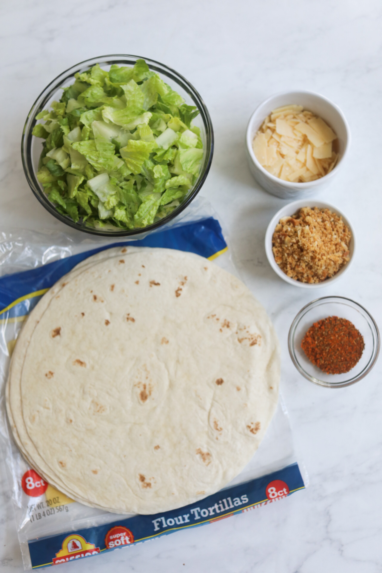 Caesar wrap ingredients. In a flat lay, large tortillas, bowl of romaine lettuce, parmesan cheese, breadcrumbs and cajun seasoning are displayed.