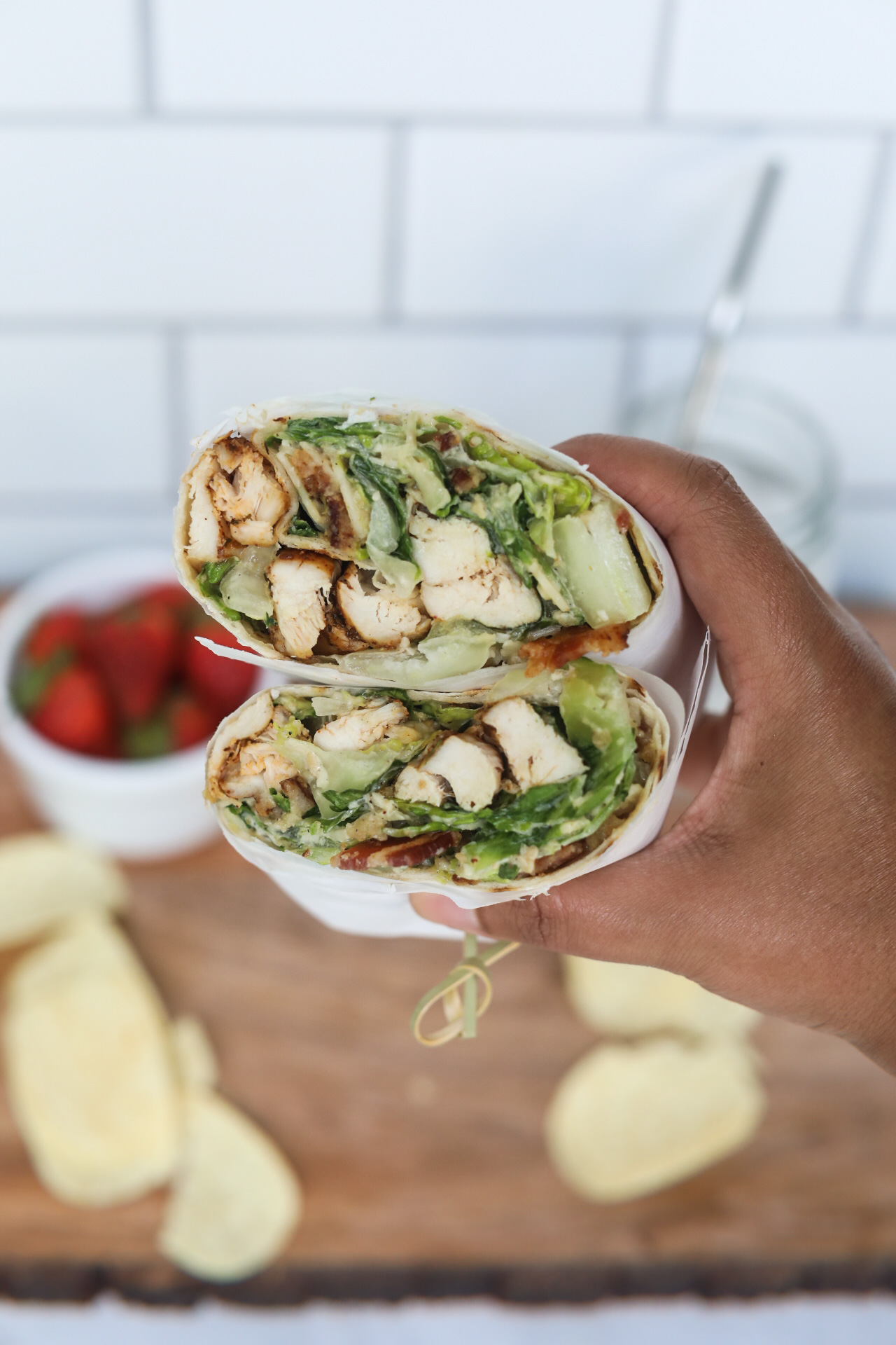 Right hand holding grilled chicken caesar wrap. The wrap is sliced open to show ingredients inside. Out of focus and for styling purposes are chips and a cup of strawberries.