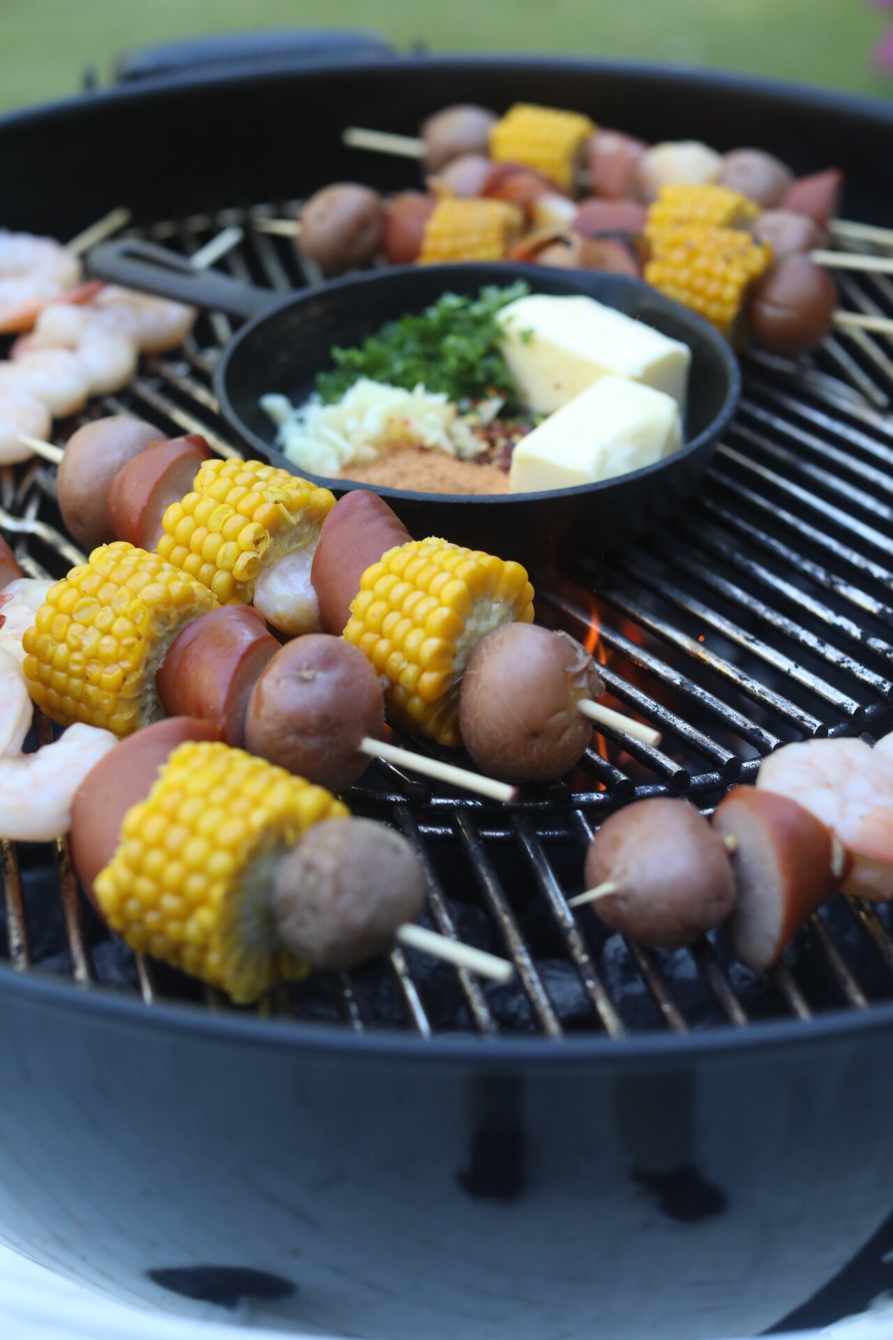 Shrimp kabobs on the grill. Skewers with potatoes, sausage, corn and shrimp on the grill. Small black cast iron skillet is placed in center with garlic butter sauce ingredients.