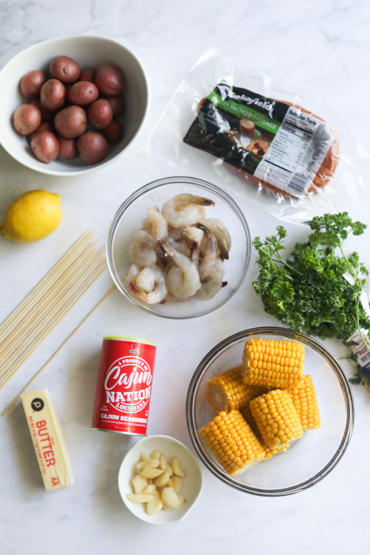 Ingredient image for grilled shrimp kabobs. On a flat lay style, small bowls with ingredients are placed. Potatoes, shrimp, corn, wooden skewers, sausage, cajun seasoning, lemon and parsley.