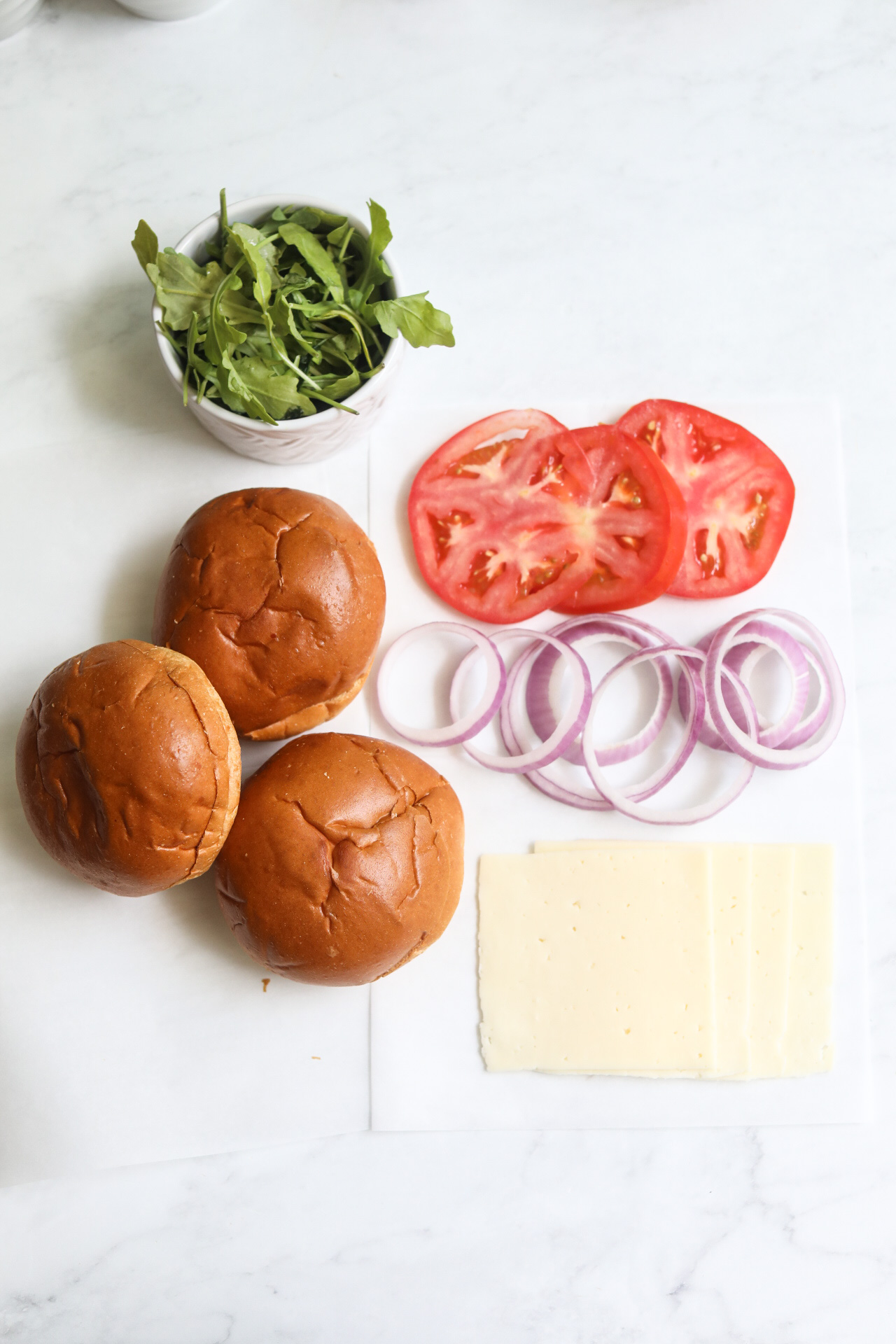 Tuna burger toppings in a flat lay. Sliced havarti cheese, brioche buns, sliced tomato, arugula and red onion.