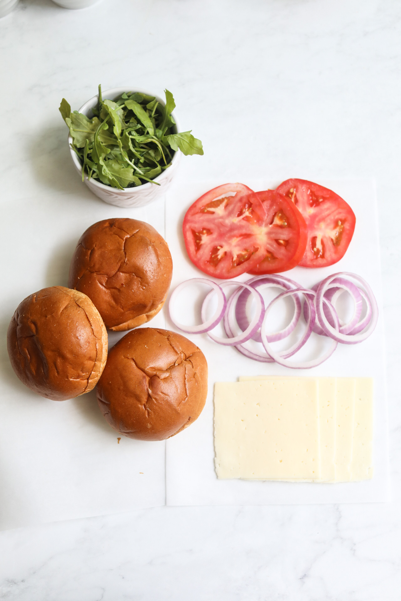 Tuna burger toppings in a flat lay. Sliced havarti cheese, brioche buns, sliced tomato, arugula and red onion. 