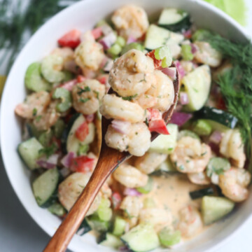 Finished recipe of shrimp salad with hand holding wooden spoon with recipe scooping out ingredients.