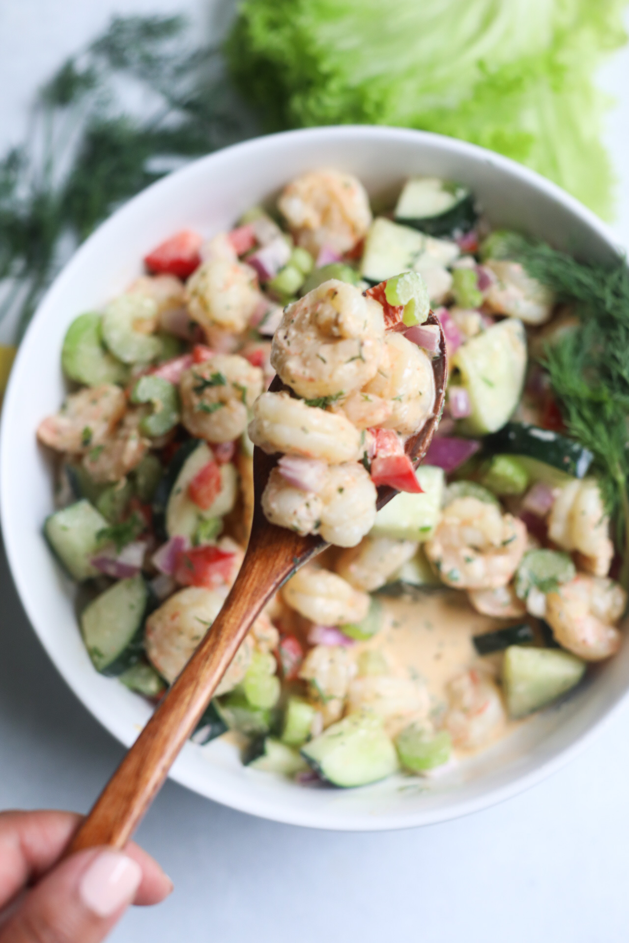 Finished recipe of shrimp salad with hand holding wooden spoon with recipe scooping out ingredients.