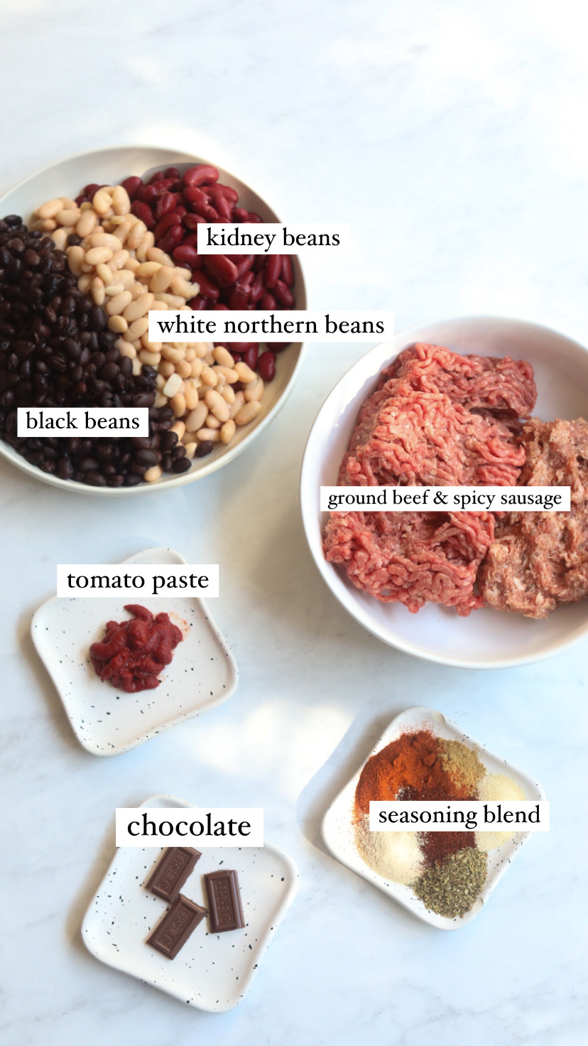 Sweet chili ingredients in a flat lay; kidney, white northern and black beans, bowl of ground beef and spicy sausage, tomato paste, chocolate and chili seasoning blend.