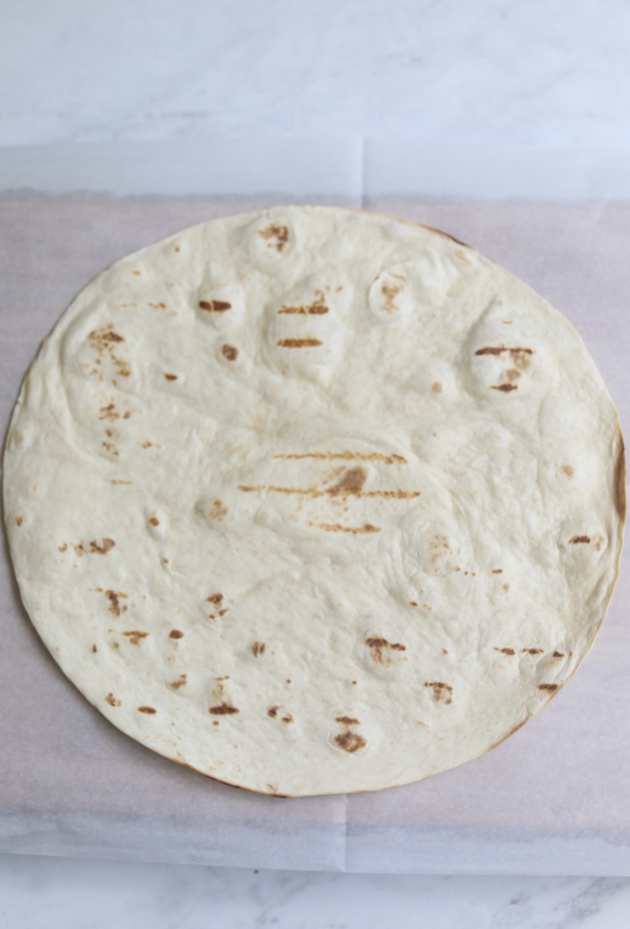 Large tortilla on wax paper ready for Grilled Chicken Caesar Wrap to be made. Image is a process shot to show steps of recipe.