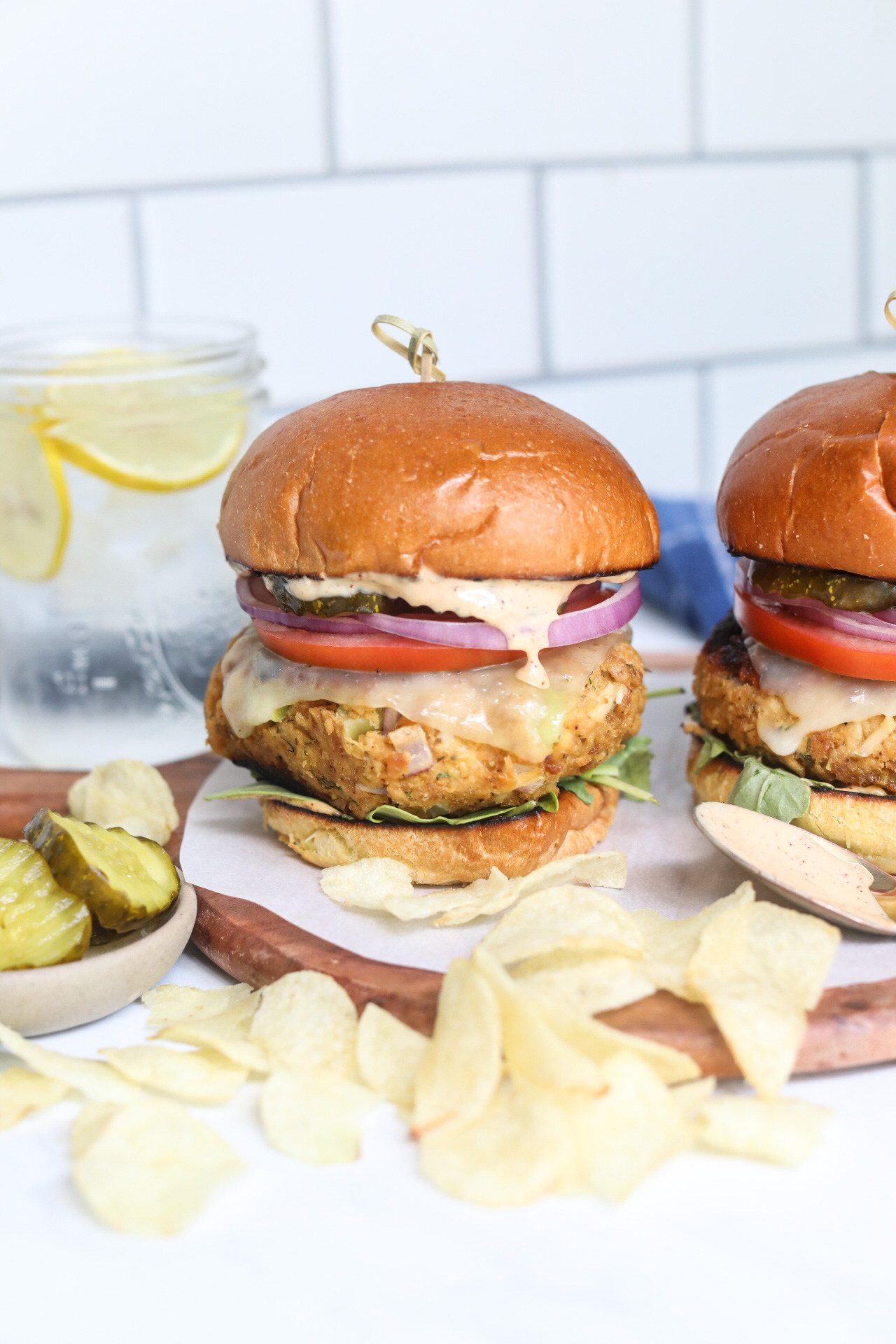 Tuna burger final shot of recipe. Burger is stacked with chips, pickles in a small bow and water glass with sliced lemon added for styling purposes.