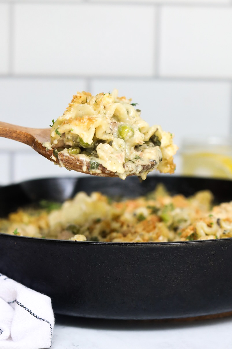 Tuna noodle casserole being dipped out of the black cast iron skillet by a wooden spoon. The image shows the depth of the recipe. 