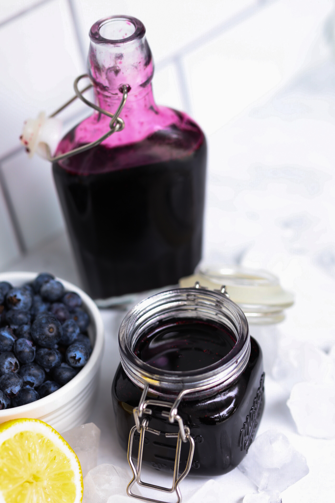 Final image of recipe in a small mason jar with lid open. Additional sauce contents are in a sauce bottle. Small white bowl with fresh blueberries, sliced lemon and ice added for styling purposes.