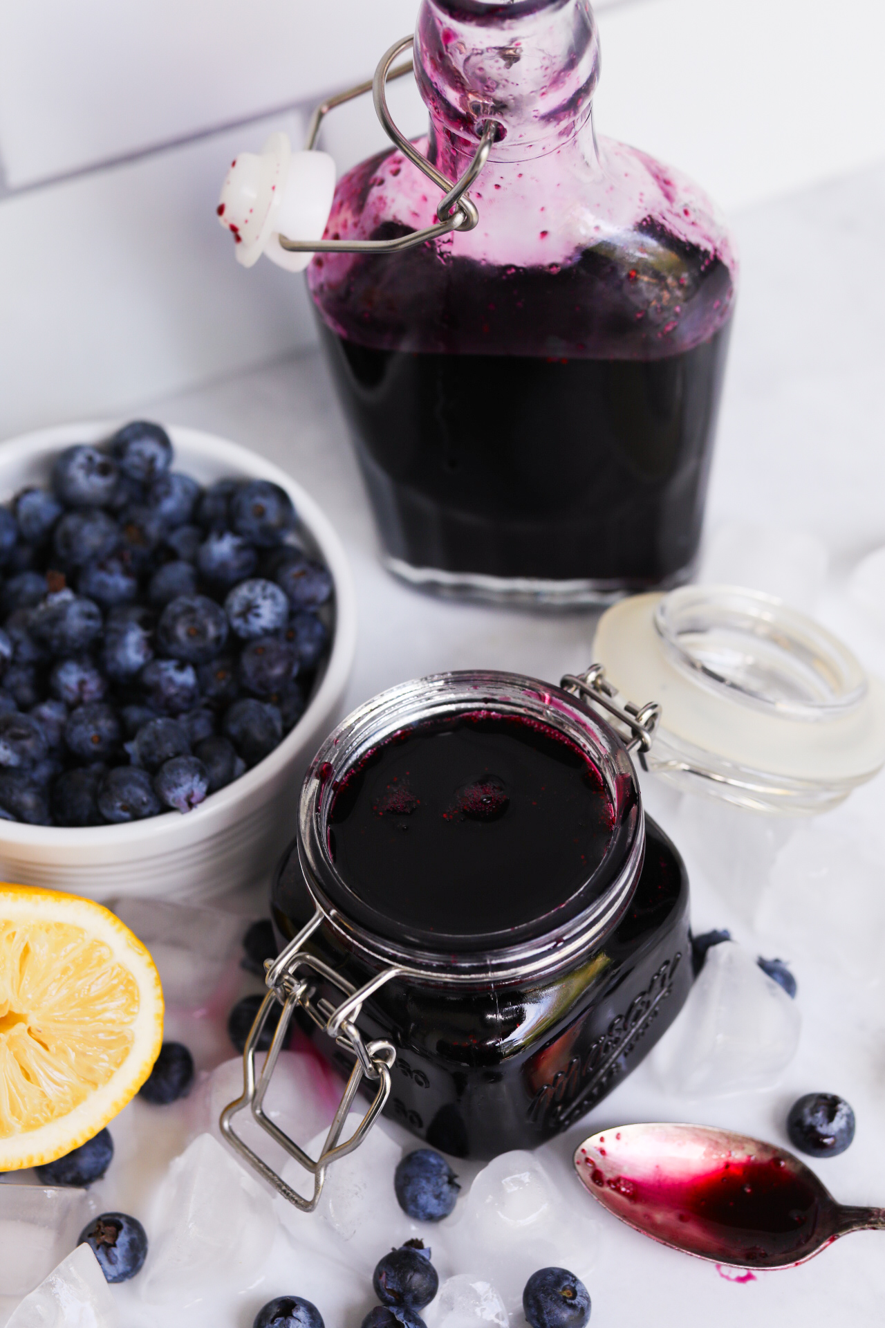 Simple syrup plated to show final recipe. Small white bowl of fresh blueberries, sliced lemon, ice chunks and scattered blueberries added for styling purposes. Mason jar and sauce jar filled with contents of blueberry simple syrup. Dainty spoon has syrup on spoon to show natural color of sauce, spoon is placed beside mason jar.