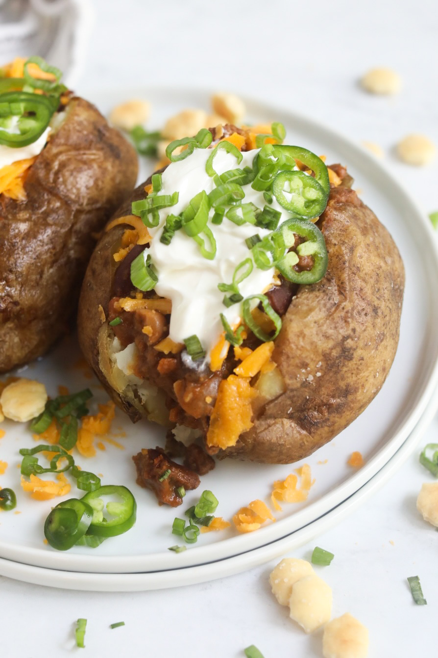 Chili stuffed baked potatoes topped with sour cream, cheddar, jalapeños, chives and cilantro. One potato is zoomed in to show contents of recipe.