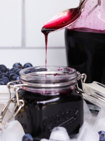 Final recipe for blueberry simple syrup. Syrup is in a small mason jar with spoon dipping out recipe. For styling purposes, small bowl of fresh blueberries, sliced lemon, ice and an additional jar of blueberry syrup.