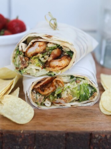 Featured image with grilled shrimp caesar wraps. Wrap is cut in half, showing contents of recipe. Chips and a small bowl of strawberries added for styling purposes.