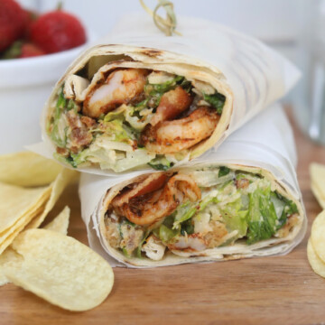 Shrimp wraps with caesar dressing cut in half and wrapped in parchment paper, stacked. Chips and a bowl of strawberries added for styling purposes.