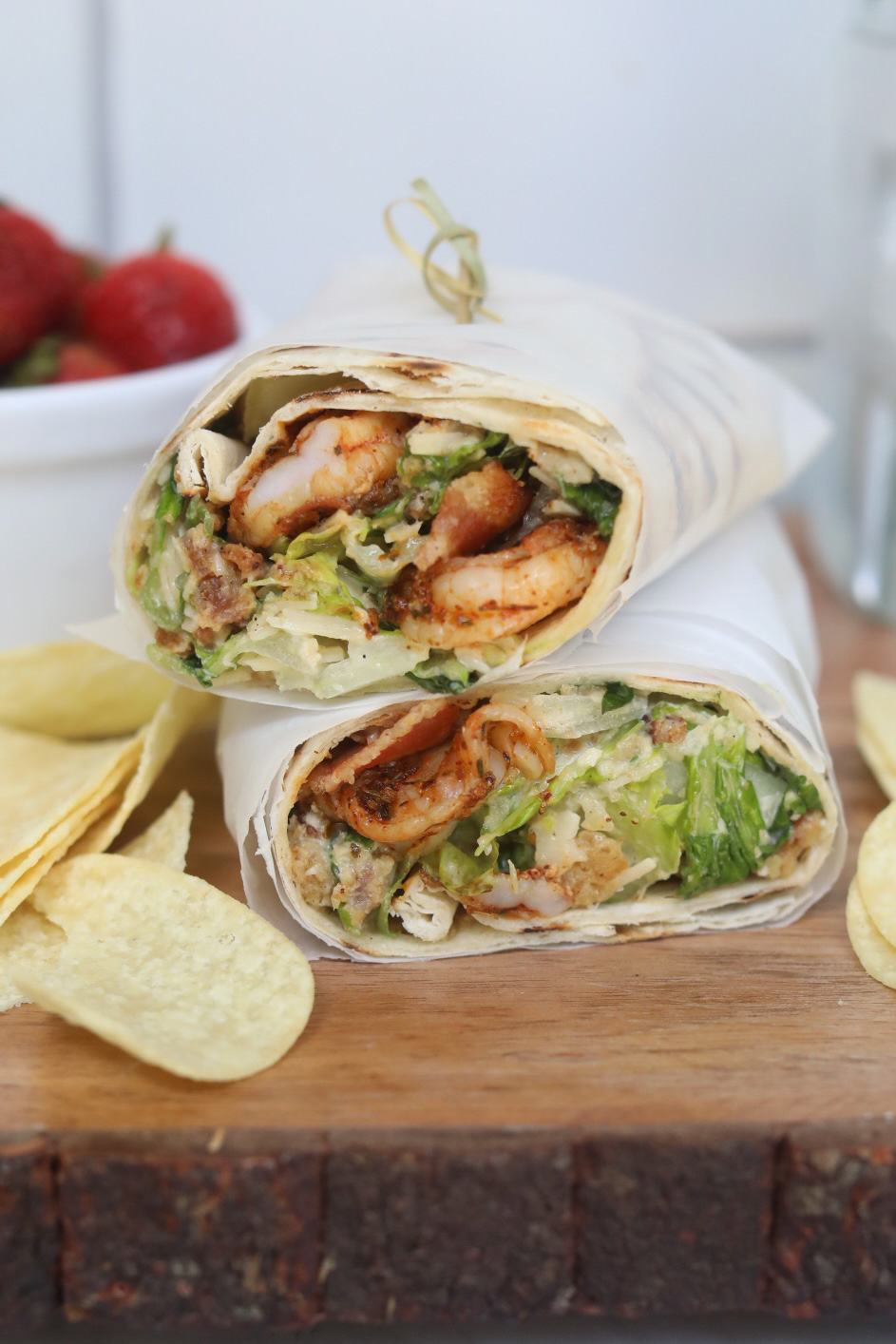 Shrimp wraps with caesar dressing cut in half and wrapped in parchment paper, stacked. Chips and a bowl of strawberries added for styling purposes.
