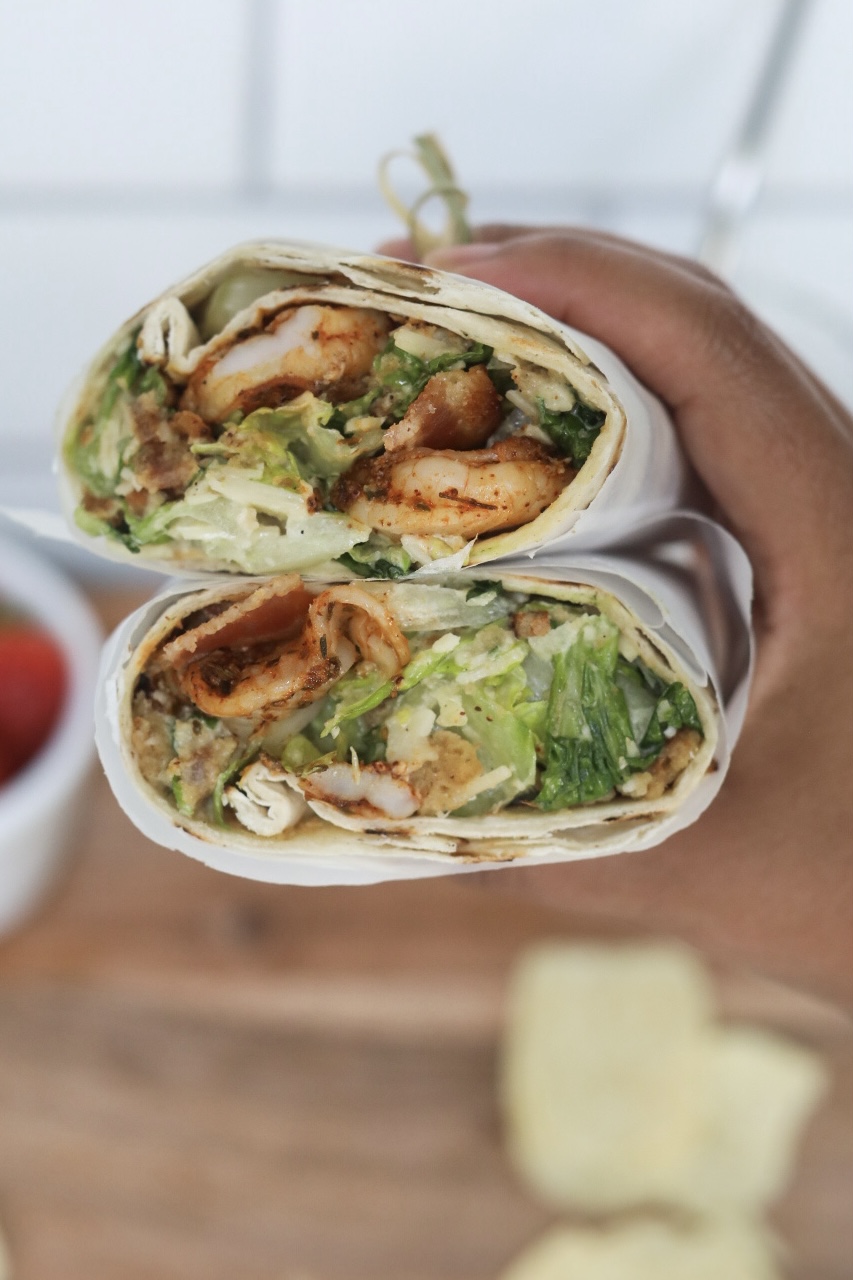 Right hand holding shrimp wraps stacked showing inside of exposed wrap with grilled shrimp, caesar salad and crispy bacon.