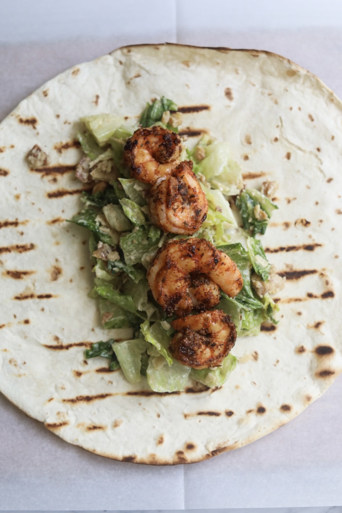 Process shot of grilled shrimp caesar wrap. Large tortilla with grill marks, caesar salad with croutons and bacon, topped with grilled cajun shrimp.