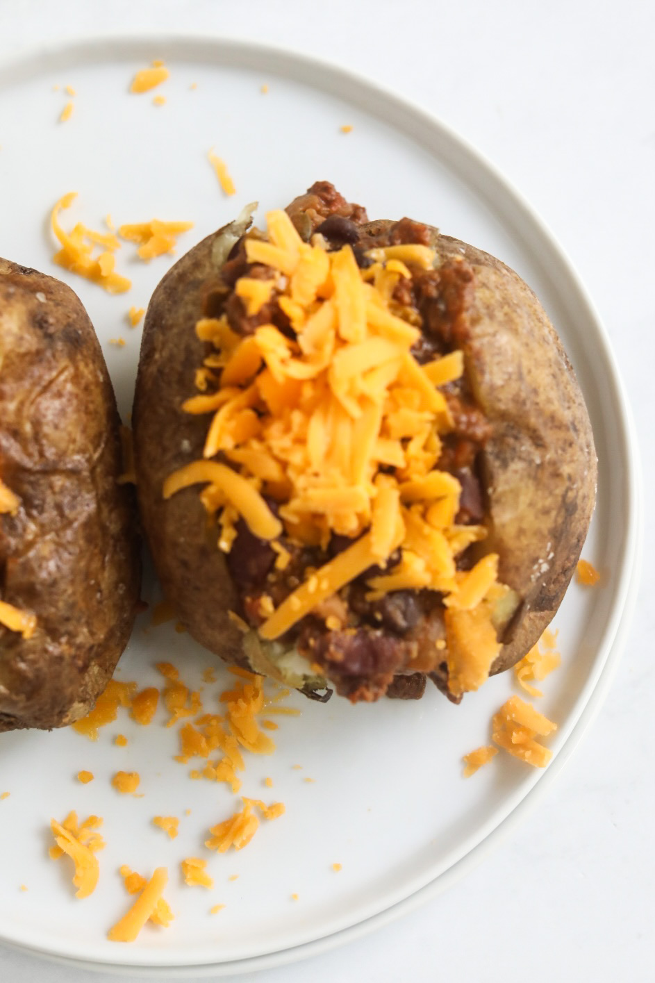 Process shot of stuffing baked potatoes. Baked potato is sliced open with chili and topped with cheddar cheese.