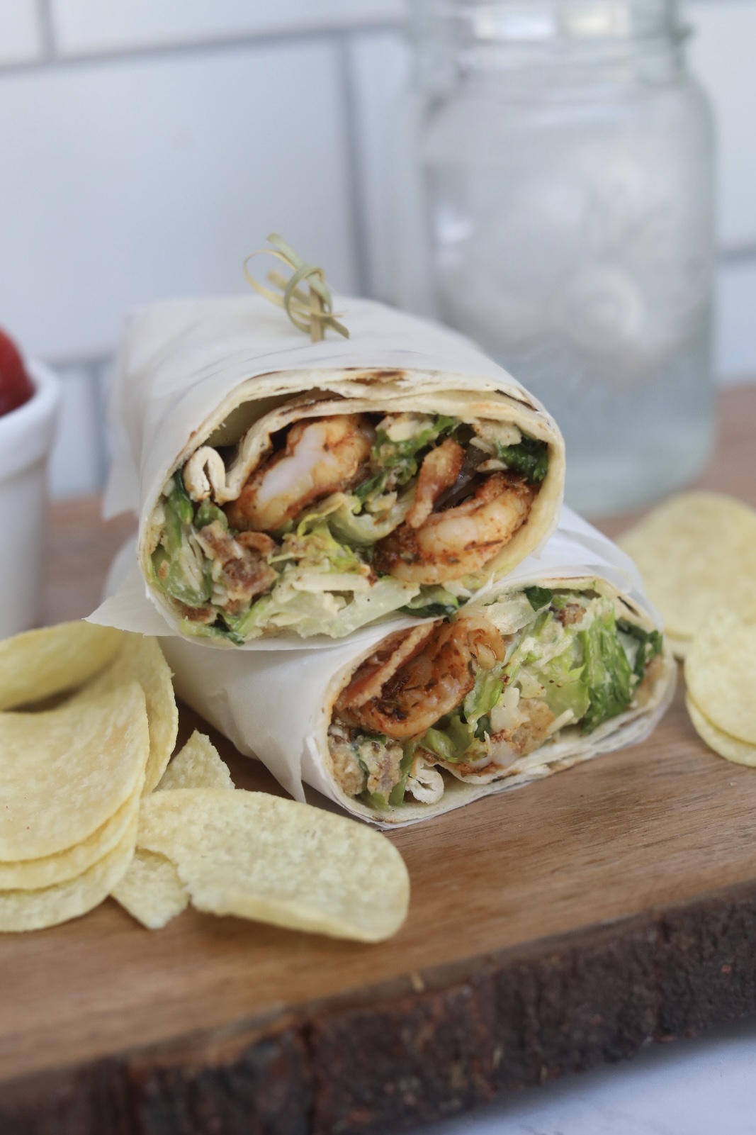 Grilled shrimp wrap sliced in half, stacked showing inside of wrap with shrimp, bacon and caesar salad. Chips, mason glass jar with water and bowl of strawberries added for styling purposes.