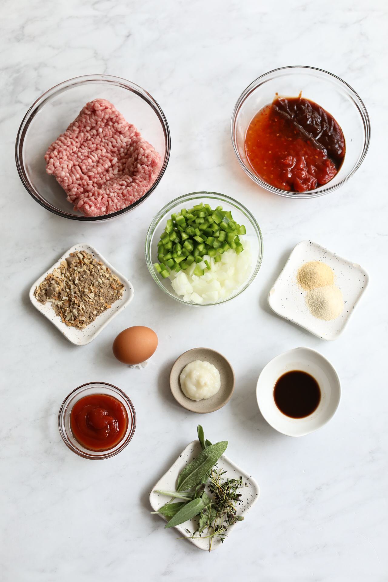 Ingredients in a flat lay with bowls of ingredients. Small bowl with ground beef and pork, meatloaf sauce, green peppers, onion, seasonings, egg, garlic, ketchup, Worcestershire sauce, fresh herbs.