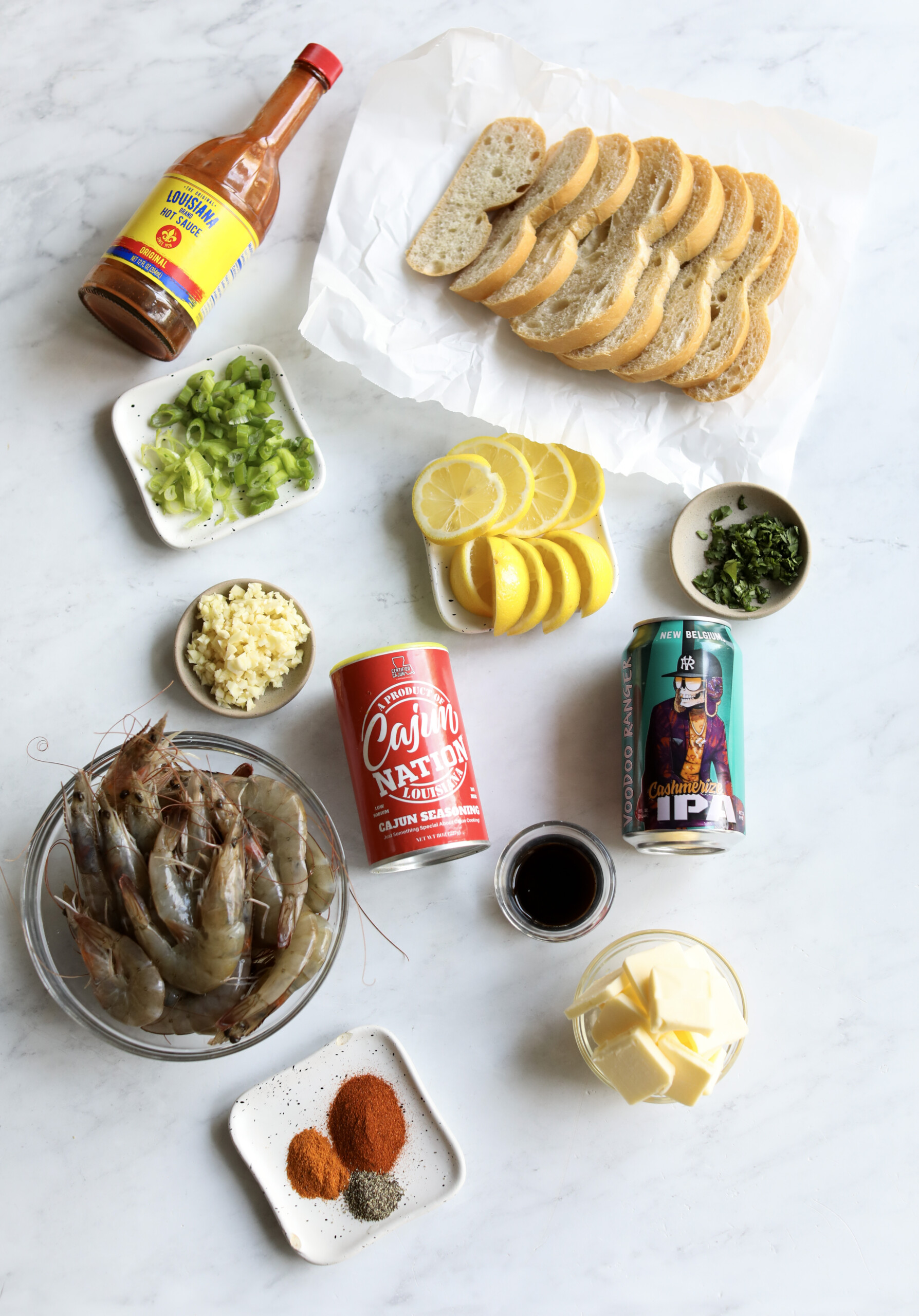 Ingredients in a flat lay for BBQ Shrimp. Bowl of head on shrimp, butter, seasonings, beer, lemons, onions and green pepper, crusty bread, Worcestershire sauce and hot sauce. 