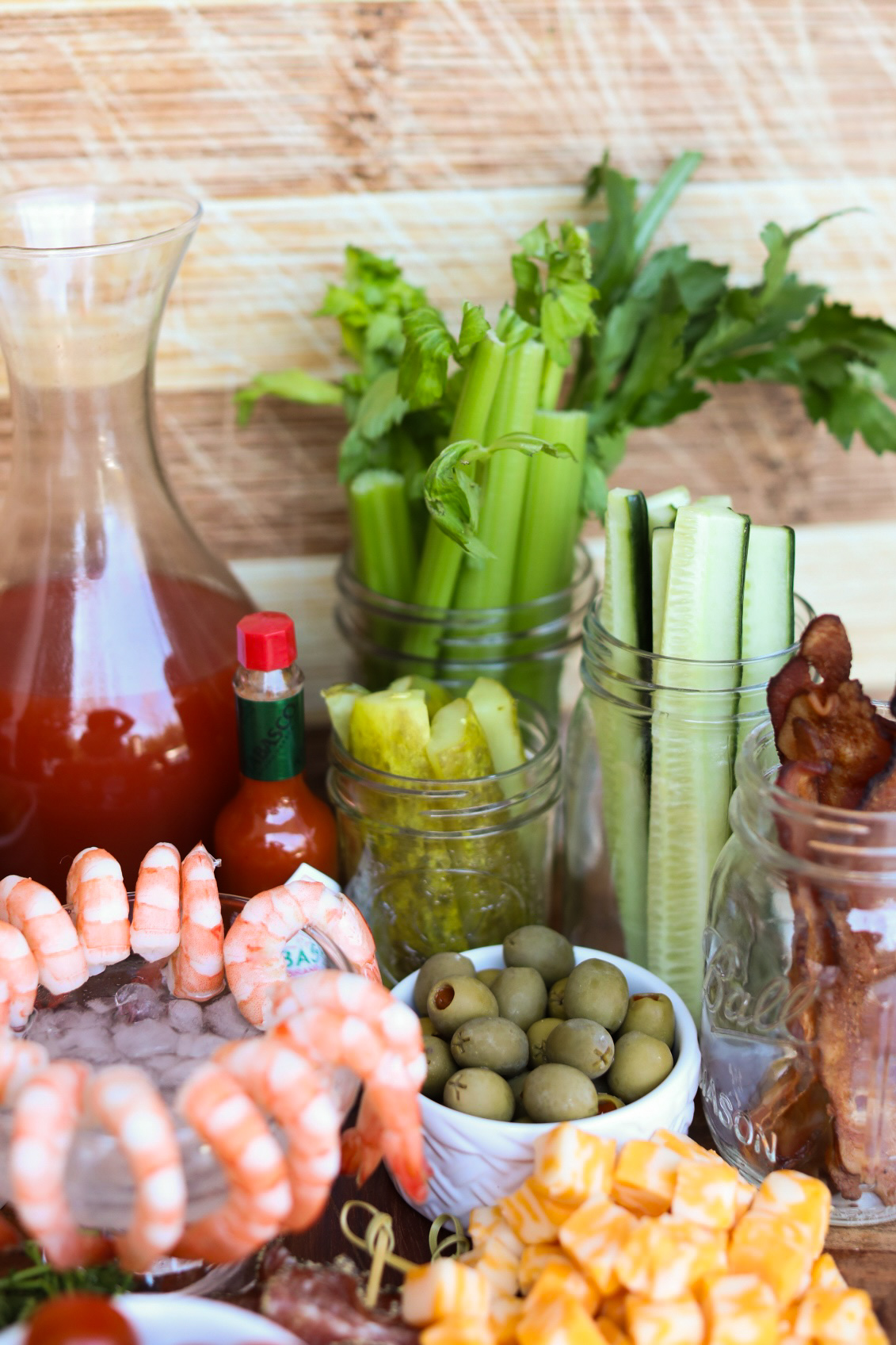 a pitcher of bloody mary beside hot sauce, shrimp cocktail, olives and bacon strips