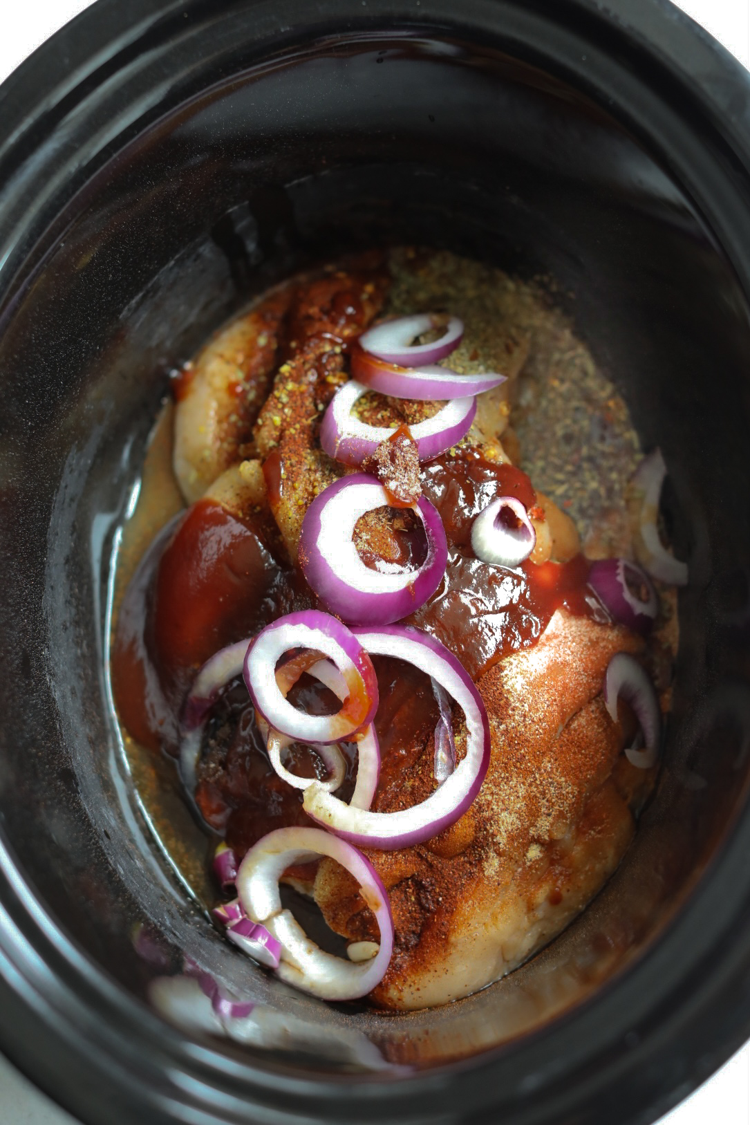 Process shot of slow cooker BBQ chicken in a black crockpot with listed ingredients before cooking process begins.