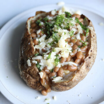 Jacket potatoes and beans served on a gray matte plate, topped with shredded cheese, chives and fresh parsley.