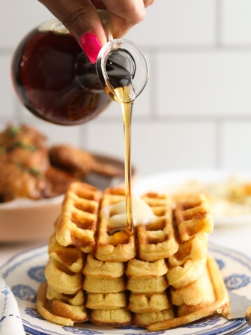 Hand holding glass syrup bottle pouring warm syrup over a stack of cornbread waffles.