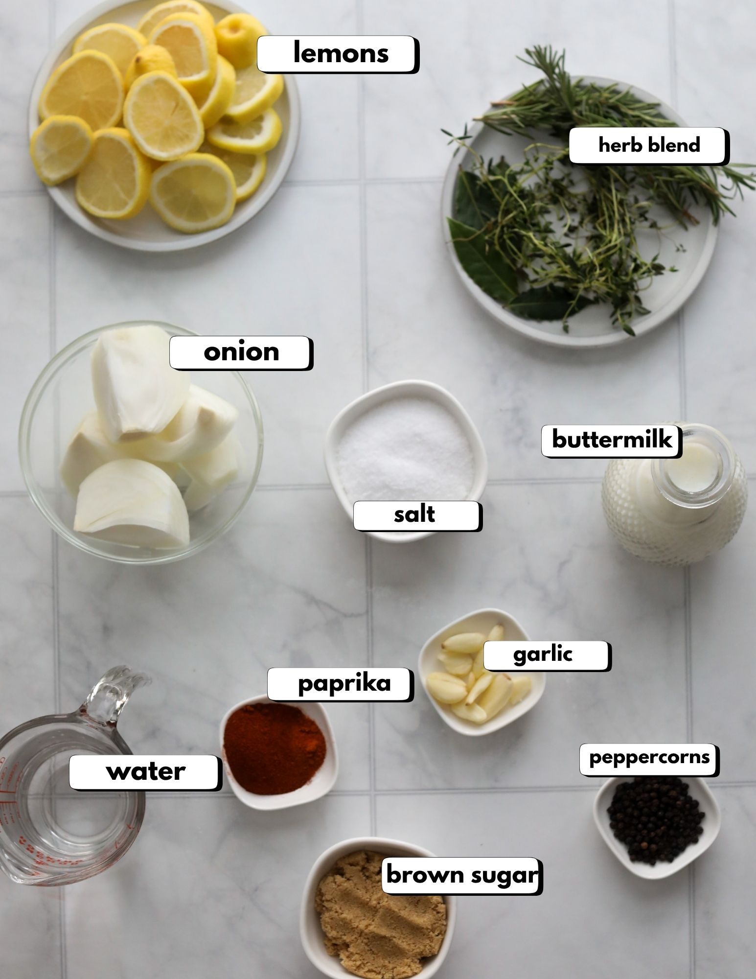 chicken brine ingredients Ingredients for chicken brine displayed on a white surface, including lemons, onion, salt, brown sugar, herbs, garlic, and spices.