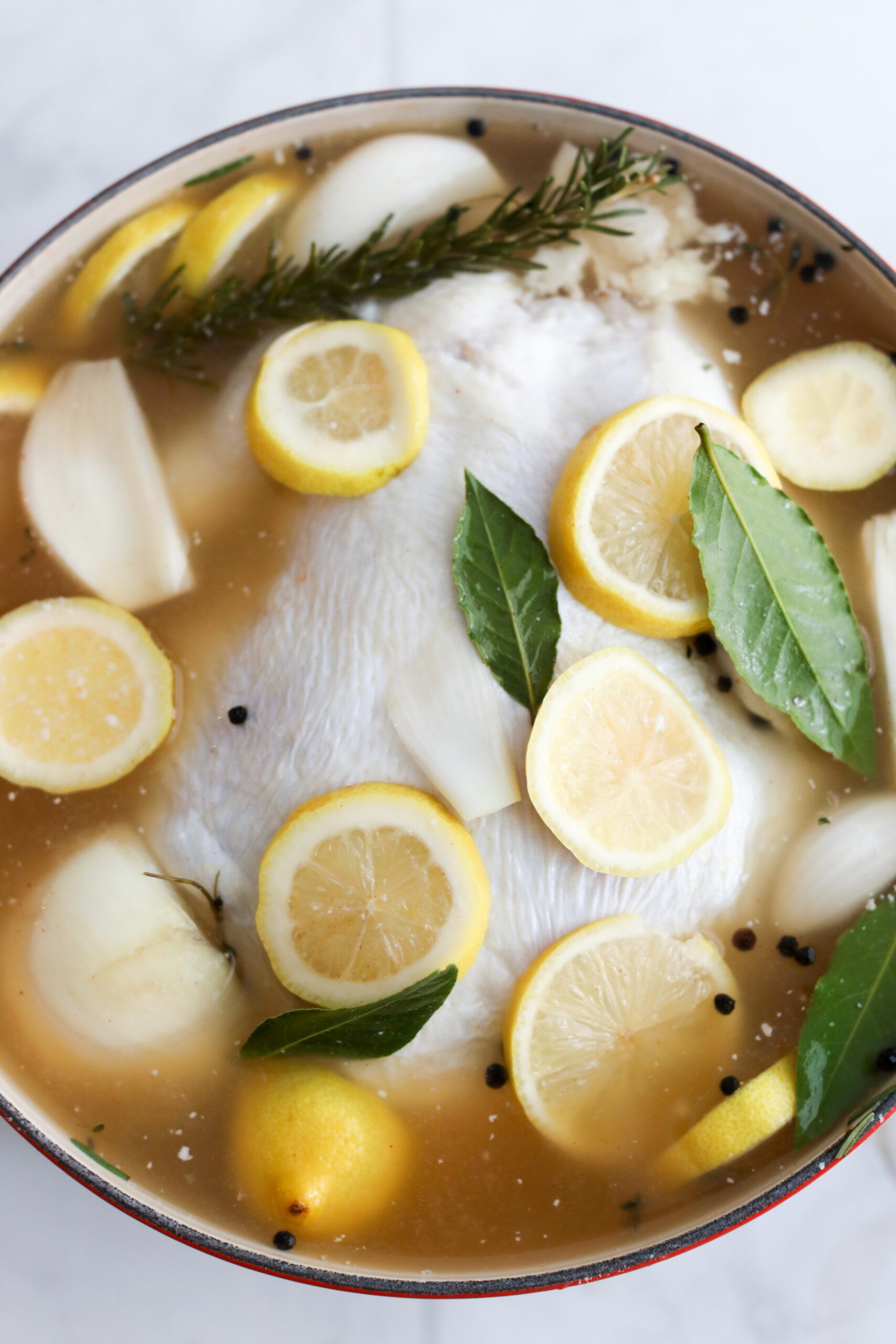 how long to brine chicken A close-up of a chicken brining in seasoned liquid with lemon slices, herbs, and black peppercorns.